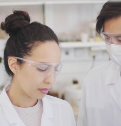 A woman in lab coat and goggles looking at another person.