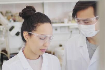 A woman in lab coat and goggles looking at another person.