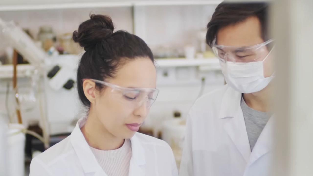 A woman in lab coat and goggles looking at another person.